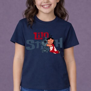 K&ouml;nnte beinhalten: Ein dunkelblaues T-Shirt mit kurzen &Auml;rmeln und einer Grafik der sich umarmenden Figuren Lilo und Stitch. Der Schriftzug Lilo & Stitch ist in roten und grauen Buchstaben &uuml;ber den Figuren aufgedruckt. Das T-Shirt hat einen Rundhalsausschnitt.