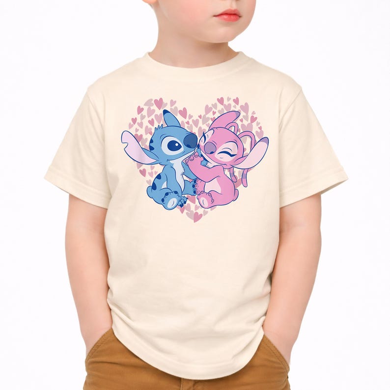 以下が含まれることがあります： クリーム色のTシャツ。青とピンクの2人の漫画のキャラクターが、小さなピンクのハートで満たされたハート型のデザインの中で抱き合っているグラフィックが特徴です。キャラクターはアニメ映画「リロ＆スティッチ」からです。