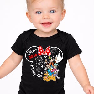 May include: A smiling baby wearing a black bodysuit with a Minnie Mouse-themed design. The design includes a red polka-dot bow, the words "Sister 2026", and Disney characters. The bodysuit is short-sleeved.