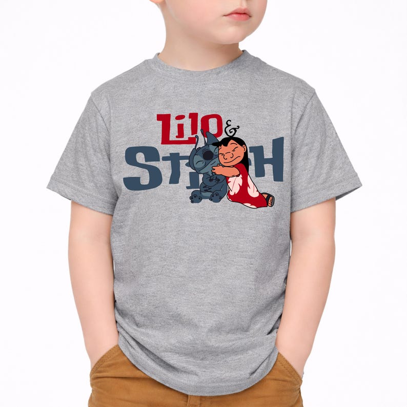 K&ouml;nnte beinhalten: Ein graumeliertes Kurzarm-T-Shirt mit einer Grafik der sich umarmenden Figuren Lilo und Stitch. Der Schriftzug "Lilo & Stitch" ist in roten und dunkelblauen Buchstaben &uuml;ber den Figuren aufgedruckt. Das T-Shirt hat einen Rundhalsausschnitt.