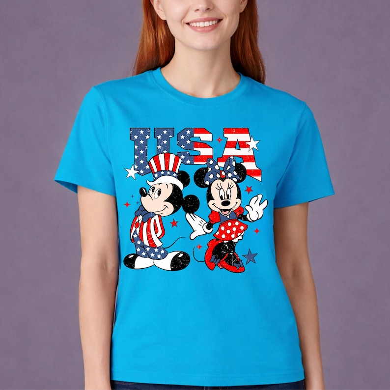 May include: A turquoise blue t-shirt featuring Mickey and Minnie Mouse in patriotic outfits. The word "USA" is displayed above the characters in red, white, and blue. The design includes stars and stripes.