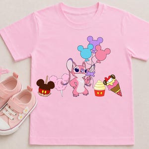 Op de afbeelding: Roze T-shirt met een stripfiguur die ballonnen vasthoudt, met illustraties van een appel met snoep, suikerspin, een cupcake en een ijshoorntje. Roze sneakers, haarclips en ringen zijn ook zichtbaar.