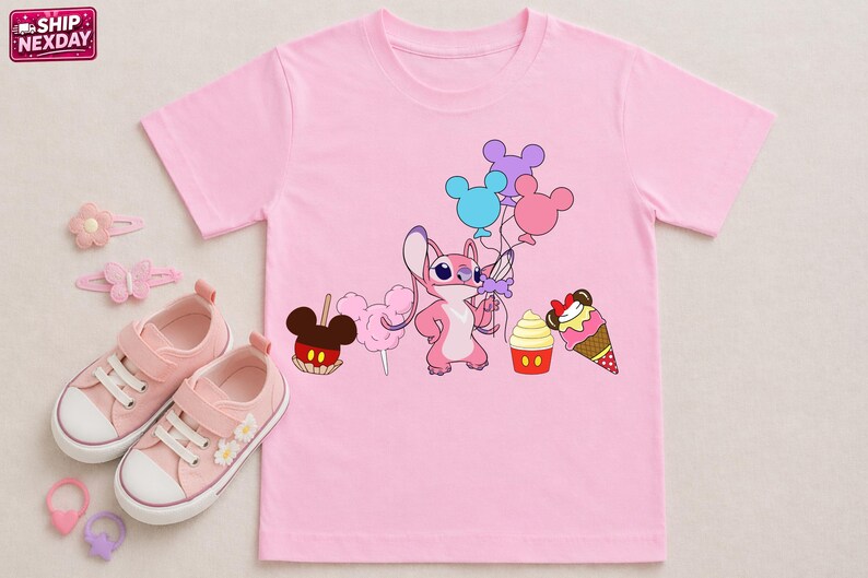 Op de afbeelding: Roze T-shirt met een stripfiguur die ballonnen vasthoudt, met illustraties van een appel met snoep, suikerspin, een cupcake en een ijshoorntje. Roze sneakers, haarclips en ringen zijn ook zichtbaar.