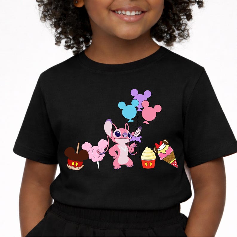 Op de afbeelding: Zwart T-shirt met een roze cartoonfiguur met een bloem en ballonnen. Het shirt heeft afbeeldingen van suikerspin, een cupcake, een ijshoorntje en een Mickey Mouse-vormige snoepappel.