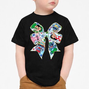 May include: Black t-shirt with a large, colourful bow design. The bow features cartoon characters on a blue floral background, with a gold outline. A fun and vibrant design.
