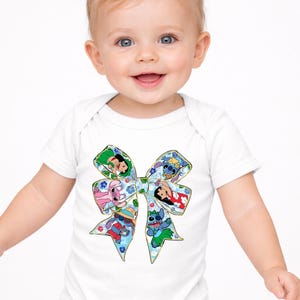 May include: White baby bodysuit with a colourful bow design featuring cartoon characters. The bow is filled with images of characters in a floral pattern. The baby is smiling and has blonde pigtails.