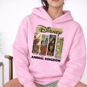 Peut inclure: Sweat &agrave; capuche rose clair avec un motif Disney. Le motif comprend le logo Disney et des images de "Le Roi Lion", "Tarzan", "Fr&egrave;re des ours" et "Le Livre de la Jungle". Le texte "ANIMAL KINGDOM" est imprim&eacute; en dessous.