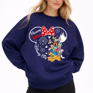 May include: Navy blue sweatshirt featuring a Disney-themed graphic. The design includes Mickey Mouse ears, a red polka-dot bow, fireworks, and various Disney characters. Text reads "Mommy 2026" and "Disney Trip".