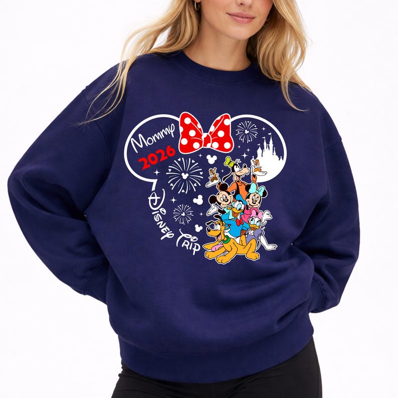 May include: Navy blue sweatshirt featuring a Disney-themed graphic. The design includes Mickey Mouse ears, a red polka-dot bow, fireworks, and various Disney characters. Text reads "Mommy 2026" and "Disney Trip".