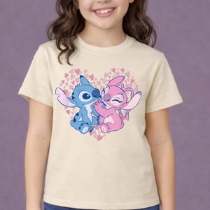 以下が含まれることがあります： ベージュ色のTシャツ。青とピンクの2人の漫画のキャラクターが、小さなピンクのハートで満たされたハート型のデザインの中で抱き合っているグラフィック。キャラクターは人気のアニメシリーズからです。
