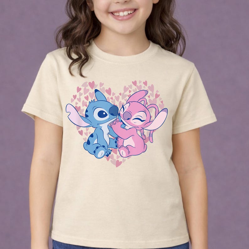 以下が含まれることがあります： ベージュ色のTシャツ。青とピンクの2人の漫画のキャラクターが、小さなピンクのハートで満たされたハート型のデザインの中で抱き合っているグラフィック。キャラクターは人気のアニメシリーズからです。