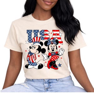 May include: Cream-coloured t-shirt featuring Mickey and Minnie Mouse in patriotic attire. The word "USA" is displayed above the characters in red and blue, with stars. Mickey wears a hat and outfit in red, white, and blue.