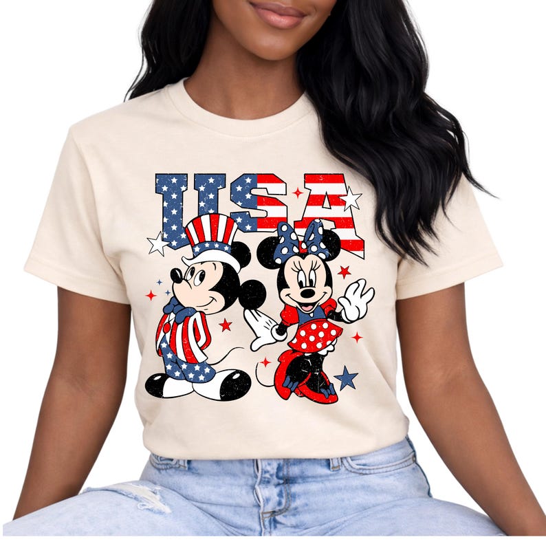 May include: Cream-coloured t-shirt featuring Mickey and Minnie Mouse in patriotic attire. The word "USA" is displayed above the characters in red and blue, with stars. Mickey wears a hat and outfit in red, white, and blue.