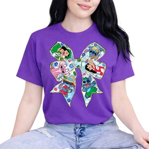 May include: A purple t-shirt with a cartoon bow design. The bow is filled with images of Stitch and Lilo characters, set against a floral background. The shirt has a crew neck and short sleeves.