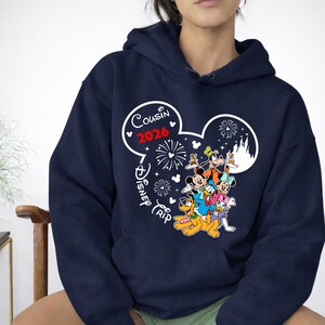 May include: Navy blue hooded sweatshirt featuring a Disney-themed graphic. The design includes Mickey Mouse ears with the text "Cousin 2026" and "Disney Trip". Various Disney characters are also depicted, including Mickey, Minnie, Donald, and Goofy.