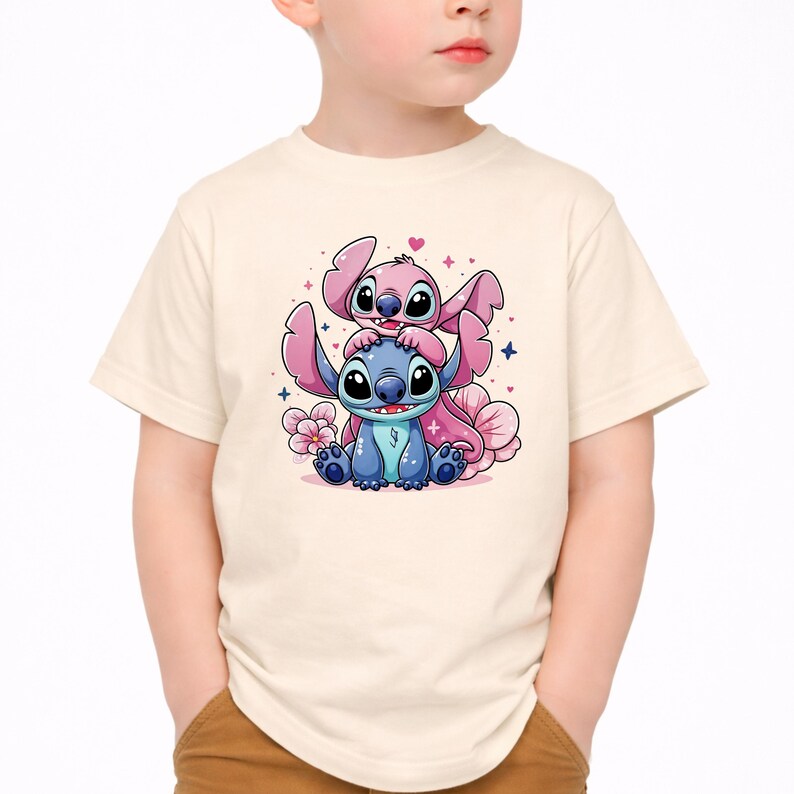 Pode incluir: Camiseta bege clara com um gr&aacute;fico colorido de dois personagens de desenho animado, um azul e outro rosa, com detalhes florais e estelares. Os personagens est&atilde;o empilhados, com o personagem rosa no topo.