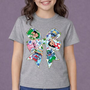 May include: A grey t-shirt with a large bow design featuring cartoon characters. The bow has a floral pattern and images of Stitch and other characters. The shirt is a short-sleeved crew neck.