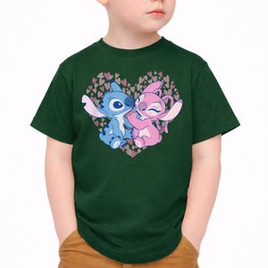以下が含まれることがあります： 濃い緑色のTシャツ。青とピンクの2人の漫画のキャラクターが、小さなハートでできたハート型のデザインの中で抱き合っているグラフィックが特徴です。キャラクターは白い背景に配置されています。