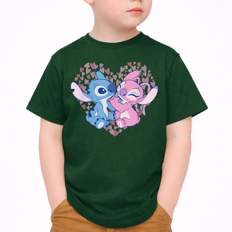以下が含まれることがあります： 濃い緑色のTシャツ。青とピンクの2人の漫画のキャラクターが、小さなハートでできたハート型のデザインの中で抱き合っているグラフィックが特徴です。キャラクターは白い背景に配置されています。