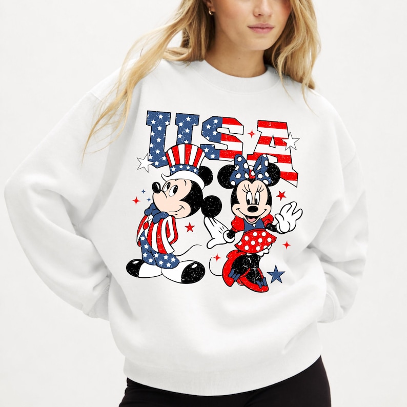 May include: White sweatshirt with a graphic of Mickey and Minnie Mouse in patriotic outfits. Mickey is in red, white, and blue, and Minnie wears a polka-dot skirt and bow. The word "USA" is printed in a distressed, star-spangled style.