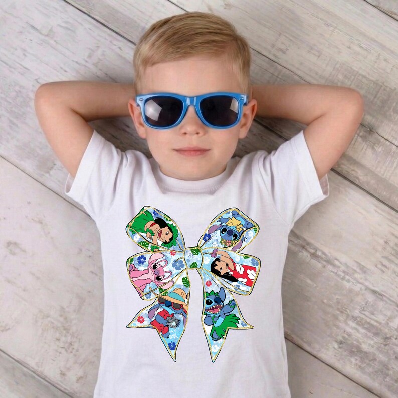 May include: White t-shirt with a large bow design featuring cartoon characters. The bow has a blue floral pattern and gold trim. The child is wearing blue sunglasses. The shirt is a children's apparel item.
