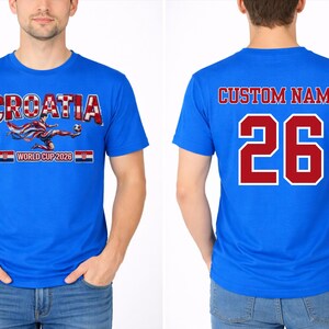 以下が含まれることがあります： 青いTシャツ。赤と白のチェック柄の「CROATIA」、サッカー選手のグラフィック、「WORLD CUP 2026」の文字。背面には「CUSTOM NAME」と赤い数字の「26」が表示されています。