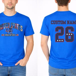 Peut inclure: T-shirt bleu roi avec l'inscription "UNITED STATES" en arc et un graphisme de football. Le devant comprend "WORLD CUP 2026". Le dos affiche "CUSTOM NAME" au-dessus du num&eacute;ro 26 avec des &eacute;toiles.