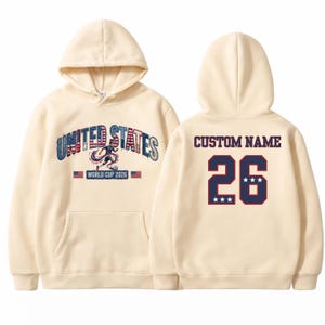 Peut inclure: Sweat &agrave; capuche cr&egrave;me avec l'inscription "UNITED STATES WORLD CUP 2026" en bleu et rouge sur le devant. Le dos affiche "CUSTOM NAME" au-dessus du num&eacute;ro "26" en bleu marine avec une bordure rouge et trois &eacute;toiles.