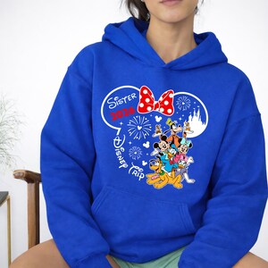 May include: Royal blue hoodie featuring a graphic of Disney characters, including Mickey and Minnie Mouse, Goofy, Donald and Daisy Duck, and Pluto. The design includes the text "Sister 2024 Disney Trip" in a white Minnie Mouse silhouette with a red polka dot bow.