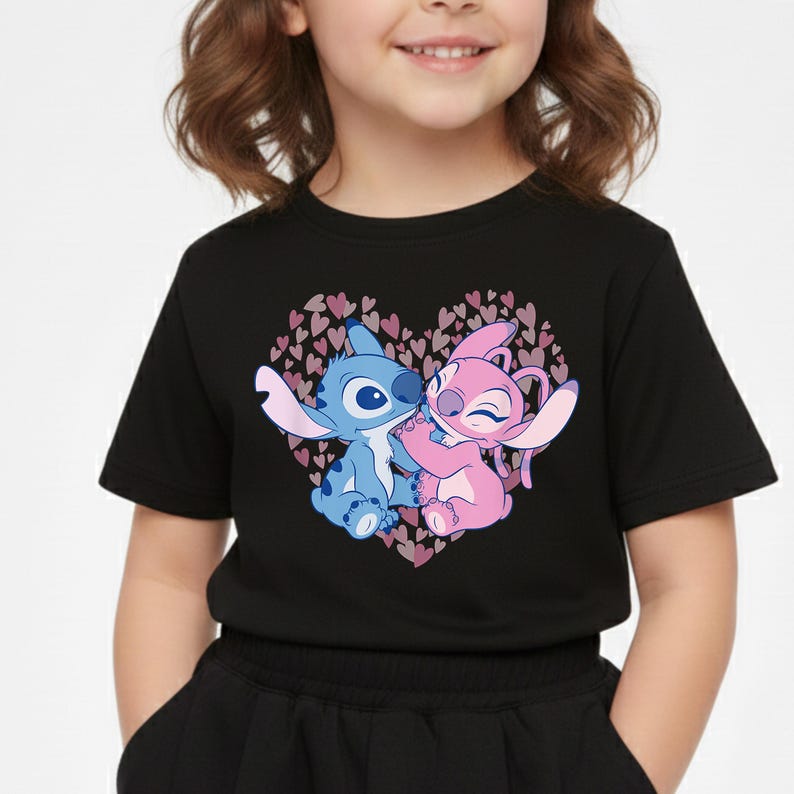 以下が含まれることがあります： 黒いTシャツ。ハート型のデザインで、漫画のキャラクターが描かれています。青いキャラクターとピンクのキャラクターが抱き合っており、小さなピンクのハートの背景があります。