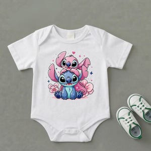 Pode incluir: Body de beb&eacute; branco com uma ilustra&ccedil;&atilde;o de desenho animado de duas personagens, uma azul e outra rosa, rodeadas por flores e cora&ccedil;&otilde;es. O conjunto &eacute; completado com sapatos de beb&eacute; brancos e verdes, oferecendo um visual alegre.
