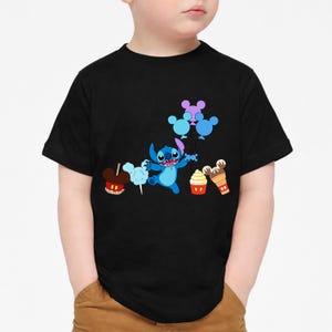 Op de afbeelding: Zwart T-shirt met een blauw stripfiguur met ballonnen en cartoon traktaties. De afbeelding bevat een Mickey Mouse-vormige karamelappel, suikerspin, een cupcake en een ijsje.