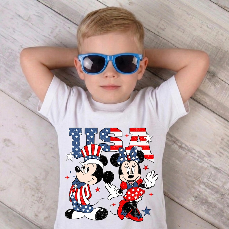 May include: A child wearing blue sunglasses and a white t-shirt. The shirt displays the word "USA" in red, white, and blue, along with illustrations of Mickey and Minnie Mouse in patriotic attire, with stars.