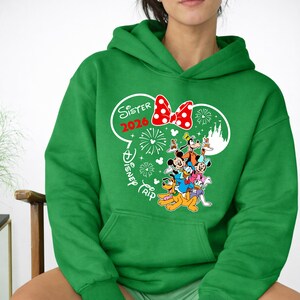 May include: A green hooded sweatshirt featuring a graphic of Disney characters, including Mickey Mouse, Minnie Mouse, and Goofy. The design includes the text "Sister 2020 Disney Trip" in a white and red color scheme. The sweatshirt has a front pocket.