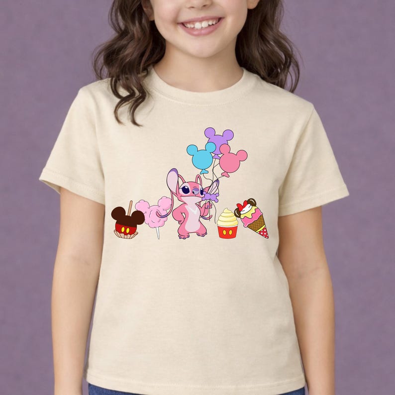 Op de afbeelding: Cr&egrave;mekleurig T-shirt met een roze cartoonfiguur met kleurrijke ballonnen. Het ontwerp bevat een snoepappel, suikerspin, een cupcake en een ijsje. Het shirt wordt door een jong persoon gedragen.