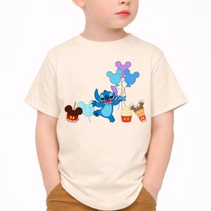 Op de afbeelding: Een cr&egrave;mekleurig T-shirt met een cartoonafbeelding van Stitch uit Lilo & Stitch. Stitch houdt ballonnen vast en is omringd door cartoonlekkernijen zoals een karamelappel, suikerspin, een cupcake en een ijsje. Het shirt is voor kinderen.