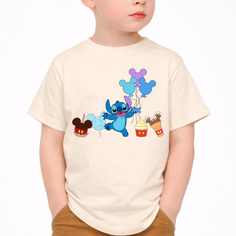 Op de afbeelding: Een cr&egrave;mekleurig T-shirt met een cartoonafbeelding van Stitch uit Lilo & Stitch. Stitch houdt ballonnen vast en is omringd door cartoonlekkernijen zoals een karamelappel, suikerspin, een cupcake en een ijsje. Het shirt is voor kinderen.