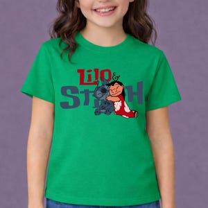 K&ouml;nnte beinhalten: Ein leuchtend gr&uuml;nes Kurzarm-T-Shirt mit einer Grafik der sich umarmenden Figuren Lilo und Stitch. Der Schriftzug Lilo & Stitch ist in Rot und Dunkelgrau auf der Brust aufgedruckt, wobei die Figuren unter dem Wort Lilo platziert sind.