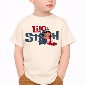 K&ouml;nnte beinhalten: Ein cremefarbenes Kurzarm-T-Shirt mit einem Grafikdruck der sich umarmenden Figuren Lilo und Stitch. Der Text Lilo & Stitch ist in roter und dunkelblauer Schrift &uuml;ber und hinter den Figuren aufgedruckt, die in ihrem klassischen Animationsstil dargestellt sind.