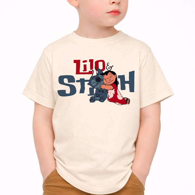 K&ouml;nnte beinhalten: Ein cremefarbenes Kurzarm-T-Shirt mit einem Grafikdruck der sich umarmenden Figuren Lilo und Stitch. Der Text Lilo & Stitch ist in roter und dunkelblauer Schrift &uuml;ber und hinter den Figuren aufgedruckt, die in ihrem klassischen Animationsstil dargestellt sind.