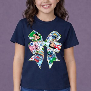 May include: A navy blue t-shirt with a large bow design featuring cartoon characters. The bow has floral patterns and gold trim. Characters include Stitch and others from the Lilo & Stitch franchise. The t-shirt is worn by a child.