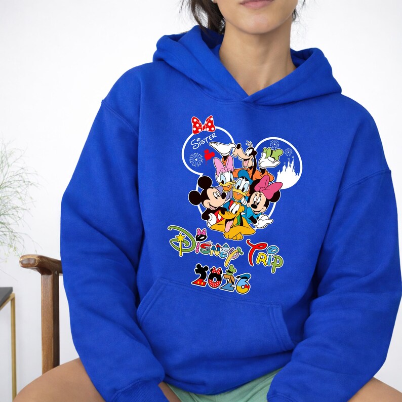May include: Royal blue hooded sweatshirt with a colourful graphic of Disney characters, including Mickey and Minnie Mouse, Donald and Daisy Duck, and Goofy. The design includes the words "Disney Trip 2026" in a playful font. A red bow is above the word "Sister".