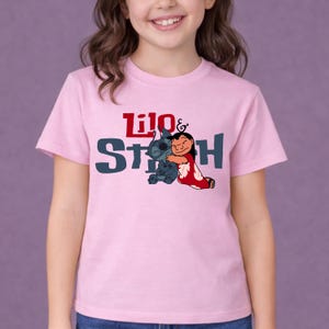 K&ouml;nnte beinhalten: Ein hellrosa T-Shirt mit kurzen &Auml;rmeln und einer Grafik der Figuren Lilo und Stitch, die sich umarmen. Der Text Lilo & Stitch ist in Rot und Dunkelblau auf der Brust aufgedruckt, zentriert &uuml;ber der Illustration der beiden Figuren.
