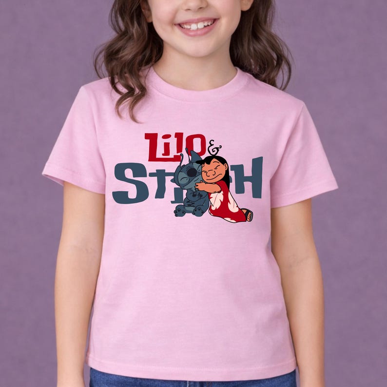 K&ouml;nnte beinhalten: Ein hellrosa T-Shirt mit kurzen &Auml;rmeln und einer Grafik der Figuren Lilo und Stitch, die sich umarmen. Der Text Lilo & Stitch ist in Rot und Dunkelblau auf der Brust aufgedruckt, zentriert &uuml;ber der Illustration der beiden Figuren.