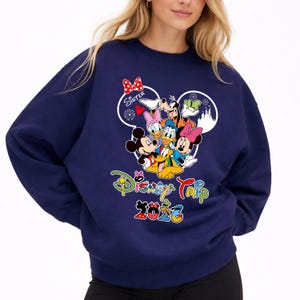 May include: Navy blue sweatshirt with a Disney Trip 2020 design. The graphic features Mickey and Minnie Mouse, Donald and Daisy Duck, Goofy, and Pluto. The word "Sister" is written above the characters in a heart-shaped design.