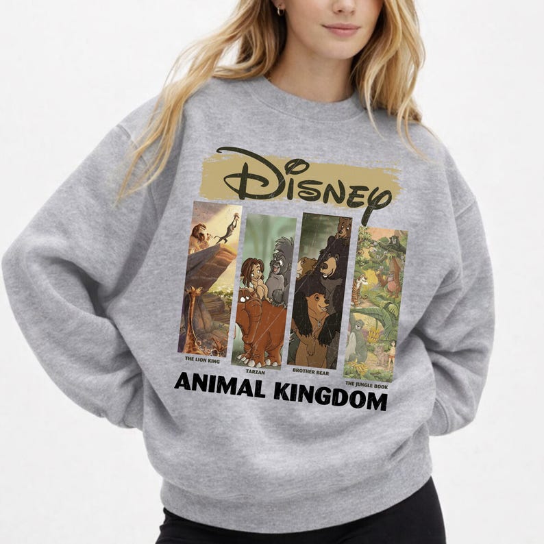 Peut inclure: Un sweat-shirt gris avec un motif Disney. Le design comprend le mot "Disney" dans une police dor&eacute;e en forme de coup de pinceau, et quatre panneaux verticaux avec des images de "Le Roi Lion", "Tarzan", "Fr&egrave;re des ours" et "Le Livre de la Jungle". Les mots "ANIMAL KINGDOM" sont imprim&eacute;s en dessous.