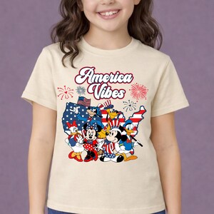 May include: A beige t-shirt featuring a graphic of Disney characters in patriotic attire, including Mickey and Minnie Mouse, Donald Duck, and Goofy, with the text "America Vibes" in red script. The design incorporates elements of the American flag.