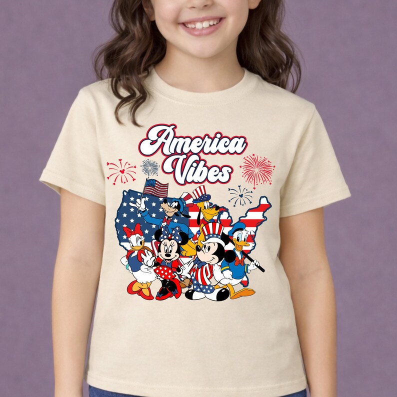 May include: A beige t-shirt featuring a graphic of Disney characters in patriotic attire, including Mickey and Minnie Mouse, Donald Duck, and Goofy, with the text "America Vibes" in red script. The design incorporates elements of the American flag.