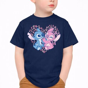 以下が含まれることがあります： ネイビーブルーのTシャツ。リロ＆スティッチのスティッチとエンジェルが、ピンクと紫のハートのハート型デザインの中で抱き合っているグラフィック。漫画のキャラクターは青とピンクの色合いです。