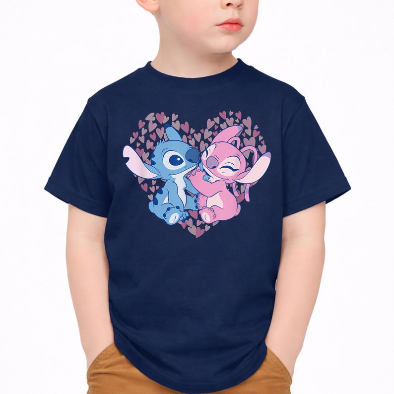 以下が含まれることがあります： ネイビーブルーのTシャツ。リロ＆スティッチのスティッチとエンジェルが、ピンクと紫のハートのハート型デザインの中で抱き合っているグラフィック。漫画のキャラクターは青とピンクの色合いです。